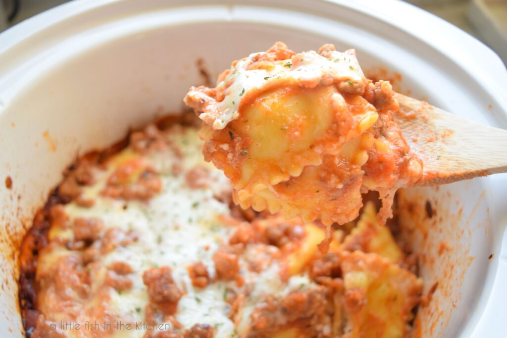 Three cheese covered cheese raviolis are being lifted by a wooden spoons. The crock with the ravioli lasagna is slightly blurred in the background. 