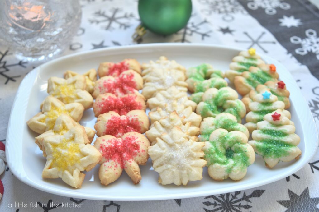 A small white platter is filled with petite spriz cookies. The cookies are shaped like Christmas trees, snow flakes, wreaths and bows. They're topped with decorative sugars that is either red, yellow or green. The platter sits on a white tea towel that is printed with gray snowflakes. A green Christmas ornaments lay beside the tin for decoration and is slightly blurred in the background.