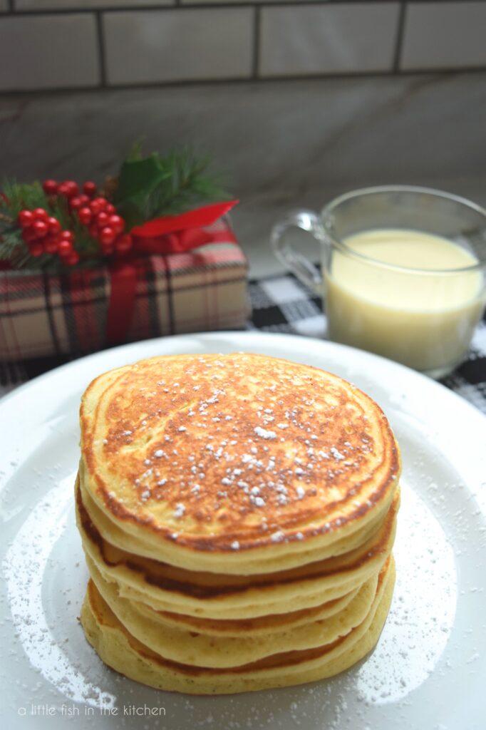 Close of up shot of the top of a stack of four egg nog pancakes. The pancakes and plate are sprinkled with powdered sugar. A small wrapped Christmas gift and a clear mug filled with egg nog are slightly blurred in the background. 