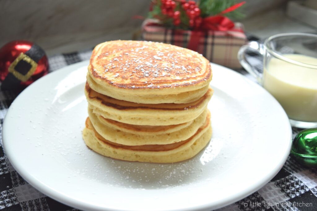 A stack of four eggnog pancakes sits on white plate that is on top of a black  and white checkered tea towel. There is a clear mug filled with eggnog, a small wrapped Christmas gift and a red and black Christmas ornament and a shiny metal jingle bell are slight blurred in the back ground.
