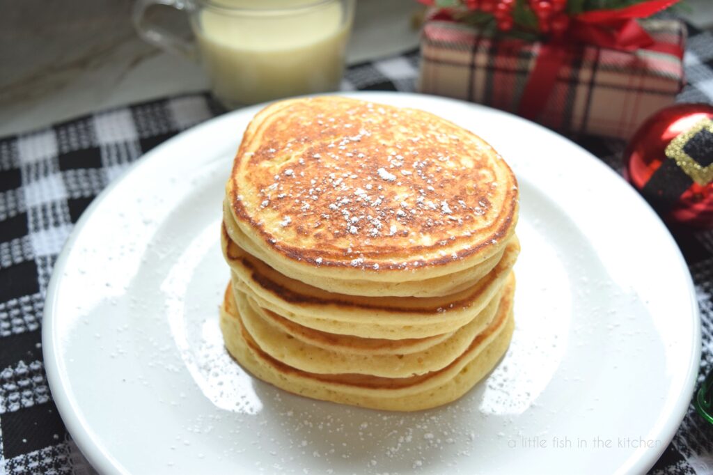 A stack of four eggnog pancakes sits on white plate that is on top of a black  and white checkered tea towel. There is a clear mug filled with eggnog, a small wrapped Christmas gift and a red and black Christmas ornament slight blurred in the back ground.