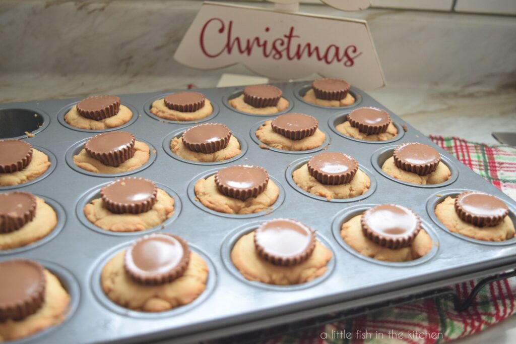 Peanut Butter Cookie Cups are pictured in the baking tin. The baking pan is on a green and red plaid tea towel. A woodent decorative sign shaped like a tree with the words Have yourself a merry little Christmas on it is slightly blurred in the background. 