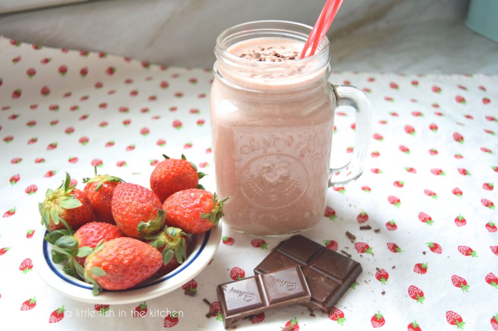 A mason jar is filled with a light pink chocolate-covered strawberry smoothie. A small bowl of fresh strawberries sits to the right of the glass and broken square of dark chocolate are stacked in front of the glass for decorations. It all sits on top of a white tea towel with a pattern of small red strawberries on it. 