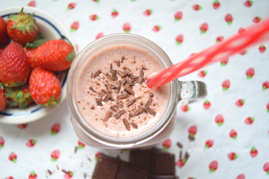 The light pink chocolate-covered strawberry smoothie from the top of the glass. Shaving of dark chocolate are on the surface and a red and white straw extends from the glass. 