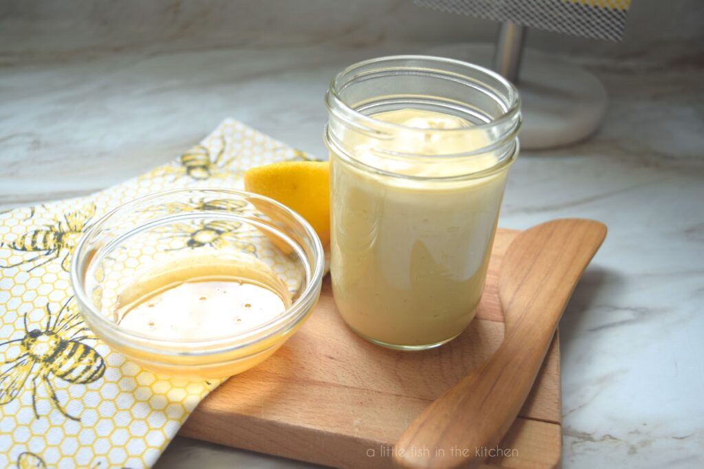 A small mason jar with honey mustard salad dressing sits atop a small wooden cutting board. The dressing is a light yellow color. A wooden butter knife, a small bowl with golden-colored honey are sitting next to the jar. A decorative tea towel with bumble bees and honey combs shapes is also draped over the cutting board handle. 