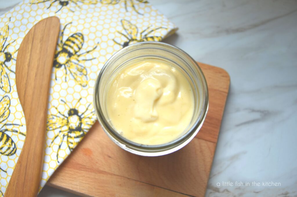 A small mason jar contains honey mustard salad dressing. It is a warm, golden yellow. A tea towel with bees and yellow honey comb pattern decoration is laying next to the jar. 