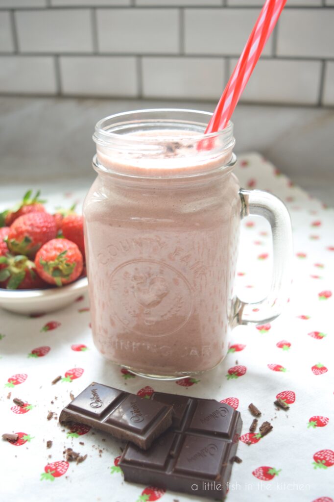A mason jar is filled with a light pink chocolate-covered strawberry smoothie. A small bowl of fresh strawberries sits to the right of the glass and broken square of dark chocolate are stacked in front of the glass for decorations. It all sits on top of a white tea towel with a pattern of small red strawberries on it.
