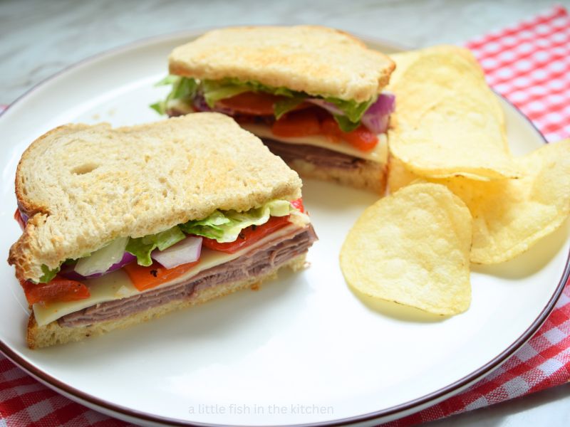 The sandwich is sliced in two exposing the colorful layers inside the bread. It's on a white plate with a small pile of potato chips beside it. The colorful layers of sliced roast beef, cheese, red peppers, red onions and lettuce are visible from the side. There is a red and white checkered tea towel lying underneath the plate