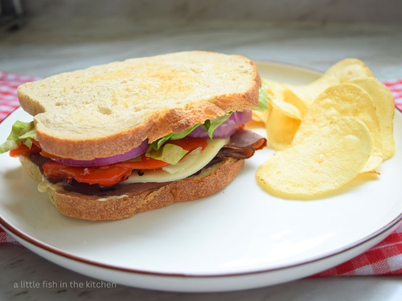 The whole Roast Beef and Red Pepper Sandwich sits on a white plate with a small pile of potato chips beside it. The colorful layers of sliced roast beef, cheese, red peppers, red onions and lettuce are visible from the side. There is a red and white checkered tea towel lying underneath the plate