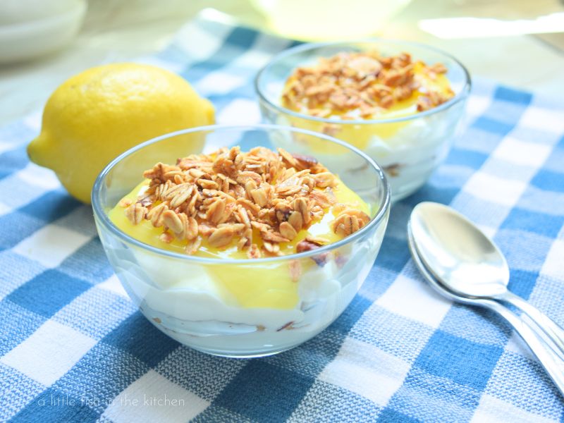 a layered lemon and yogurt parfait is ready to serve. the layers of yogurt, granola and lemon topping are visible from the side of the clear dessert bowl. 