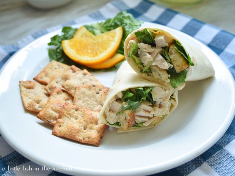 A wrap sandwich is cut in half and bits of green romaine lettuce, croutons, and chicken are visible inside the tortilla. Pita chips an slices of a fresh orange are on the plate too. 