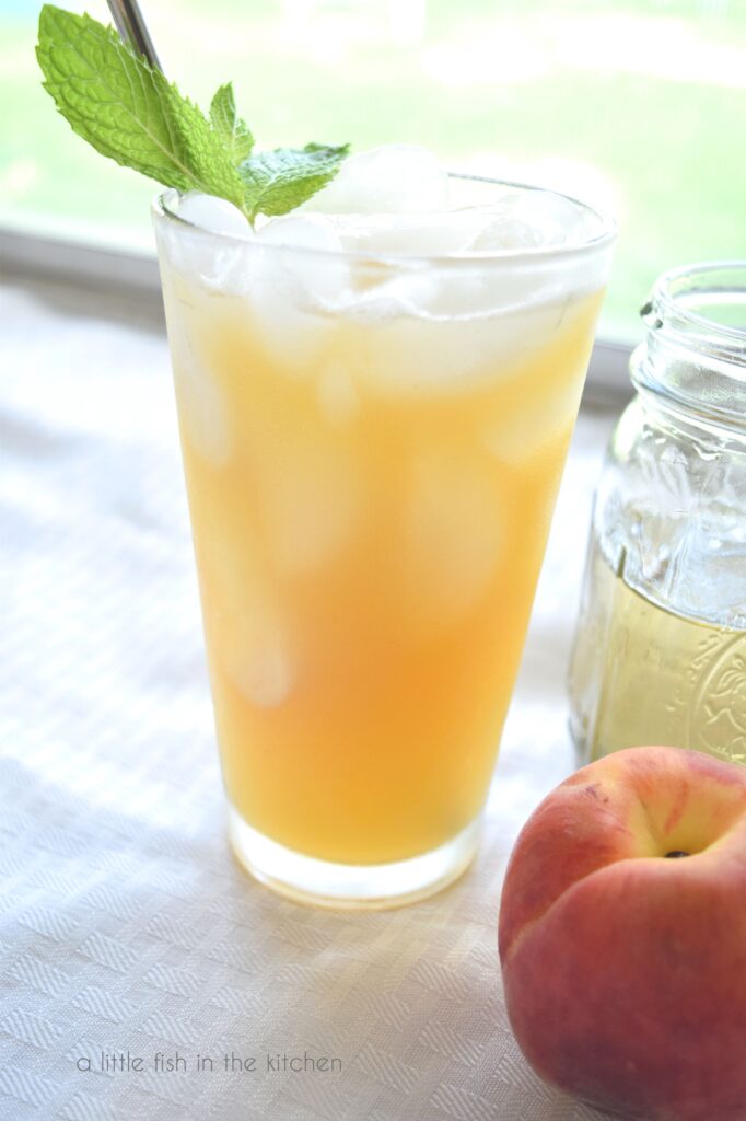 A glass with a lovely peach colored drink sits on a white tablecloth. Ice cubes are visible from the side and the drink looks cold and inviting next to a window.