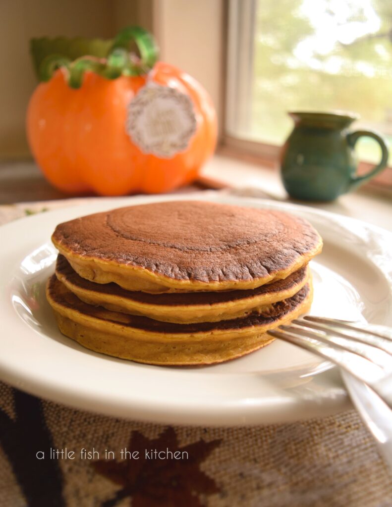 A stack of three amber-colored pancakes sit in the center of a white plate. A decorative orange glass pumpkin is slightly blurred in the background.