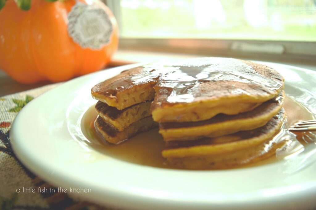 A stack of three pancakes has a been sliced and the fluffy inside of the pancakes is visible. The pancakes are in a white plate in a small pool of maple syrup. 