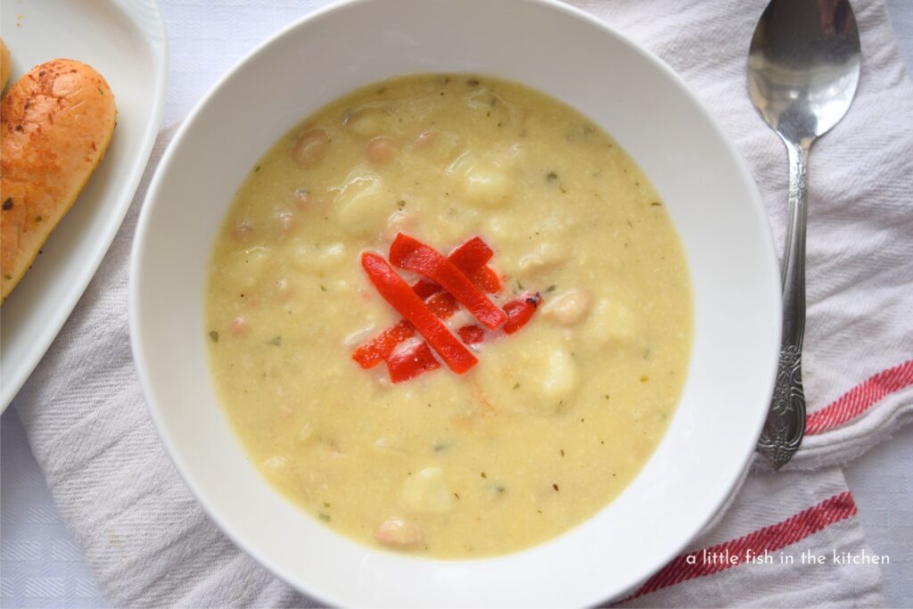 Golden-colored, creamy chowder sits in a white bowl atop a white tea towel with a red stripe. A spoon and a plate with bread sticks is on the side. 