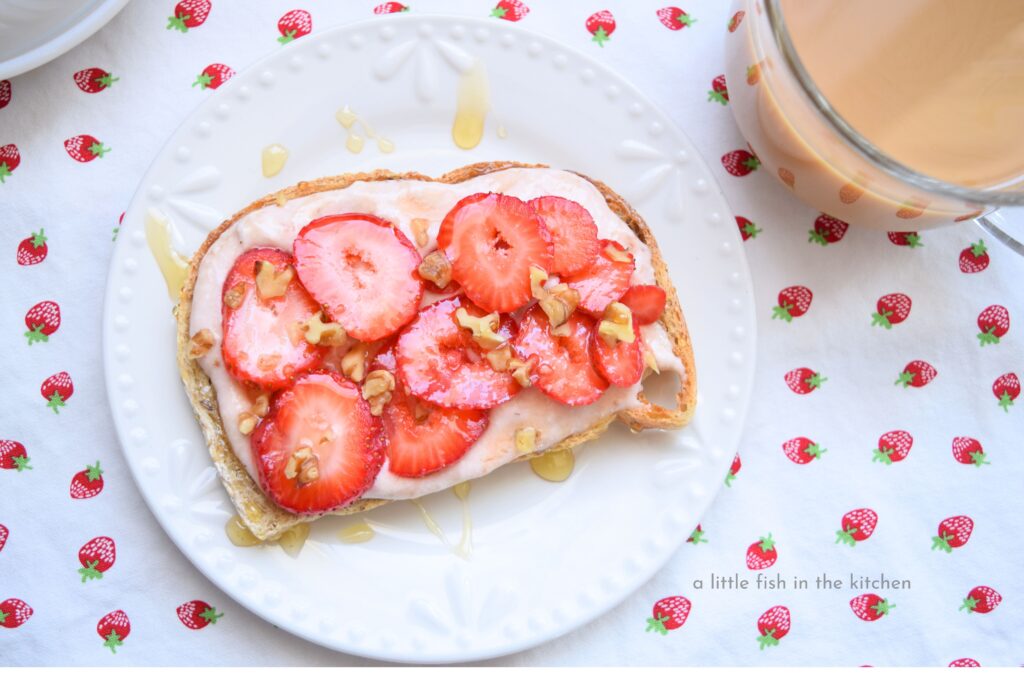 A slice of loaded strawberry cheesecake toast is ready to serve. The toast is on a white dessert plate, and served with a fresh cup of coffee.