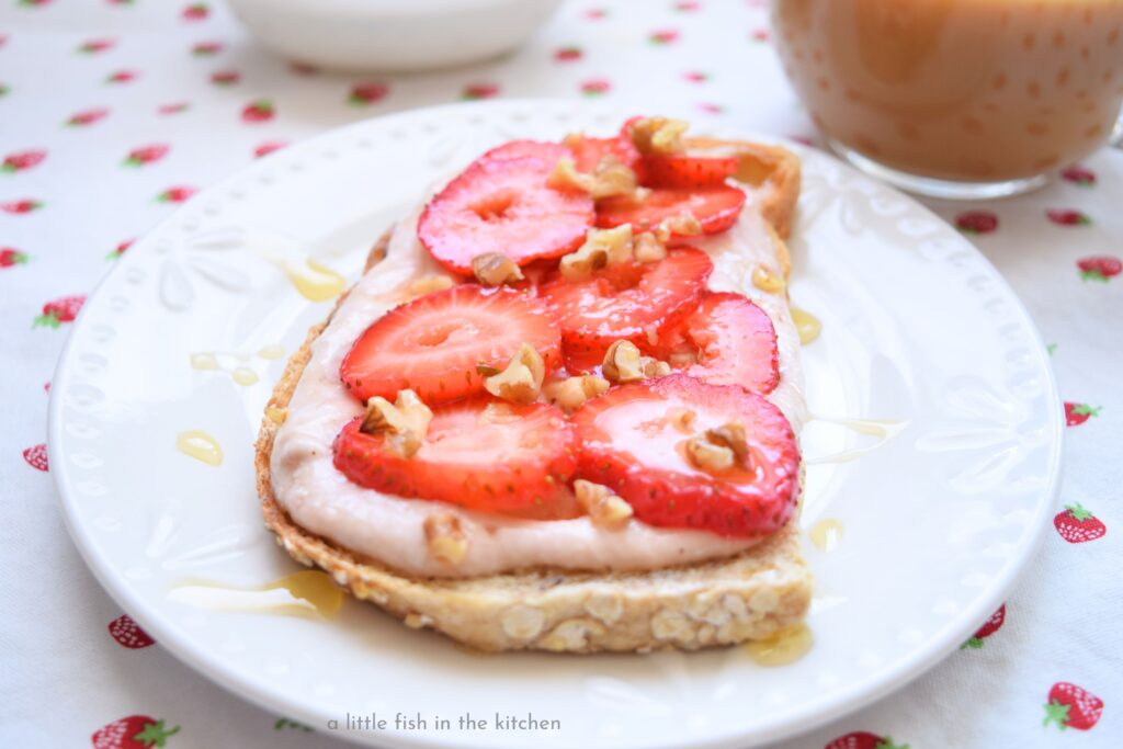 A piece of whole grain toast is topped with layers of strawberry-flavored cream cheese, slices of ripe fresh strawberries, chopped pecans and drizzled of golden local honey.
