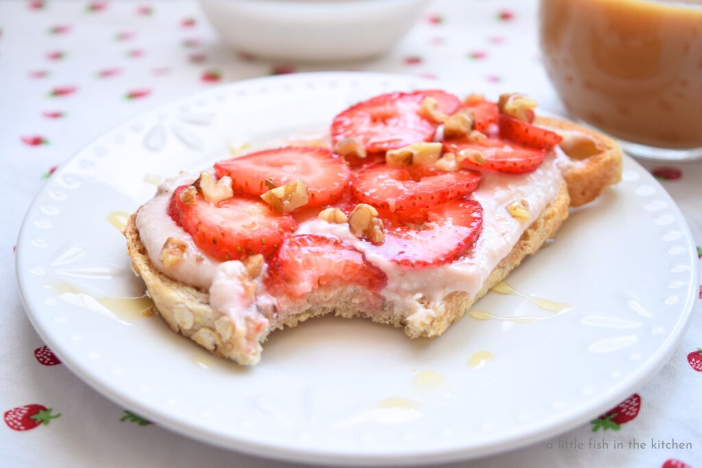 A piece of whole grain toast is topped with layers of strawberry-flavored cream cheese, slices of ripe fresh strawberries, chopped pecans and drizzled of golden local honey.