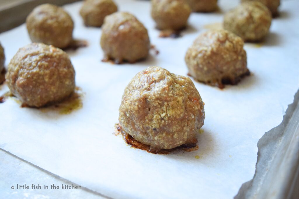 Meatballs are hot and fresh out of the oven. They are on a sheet pan lined with parchment paper. 