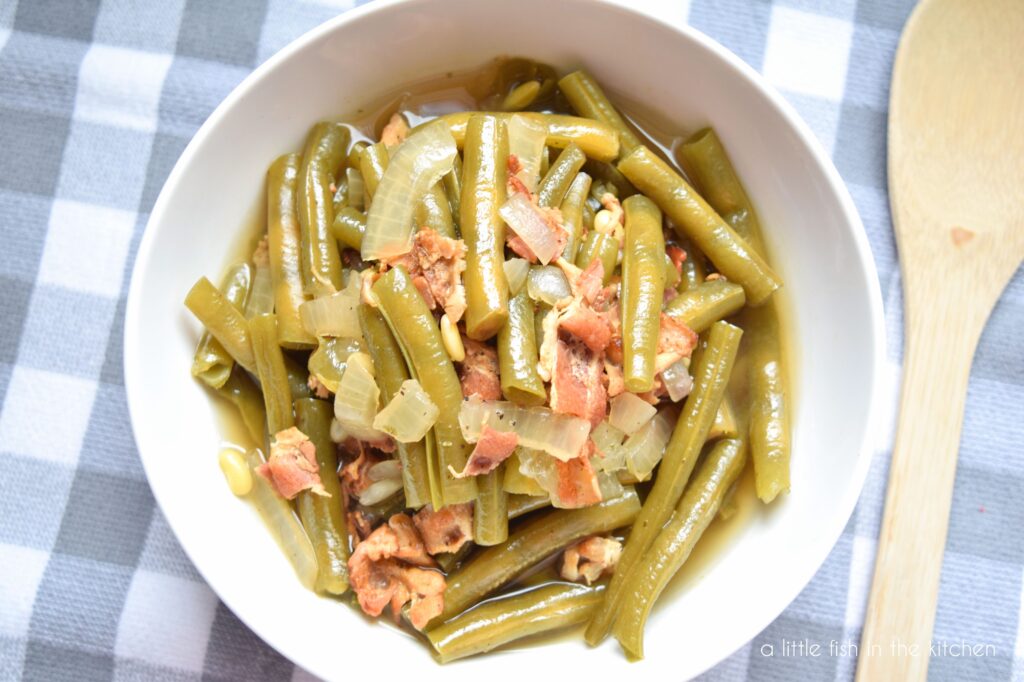 Tender-looking green beans with bits of cooked bacon and onion are ready to serve in a white bowl. A wooden spoon sits beside the bowl on a grey and white checkered tea towel. 
