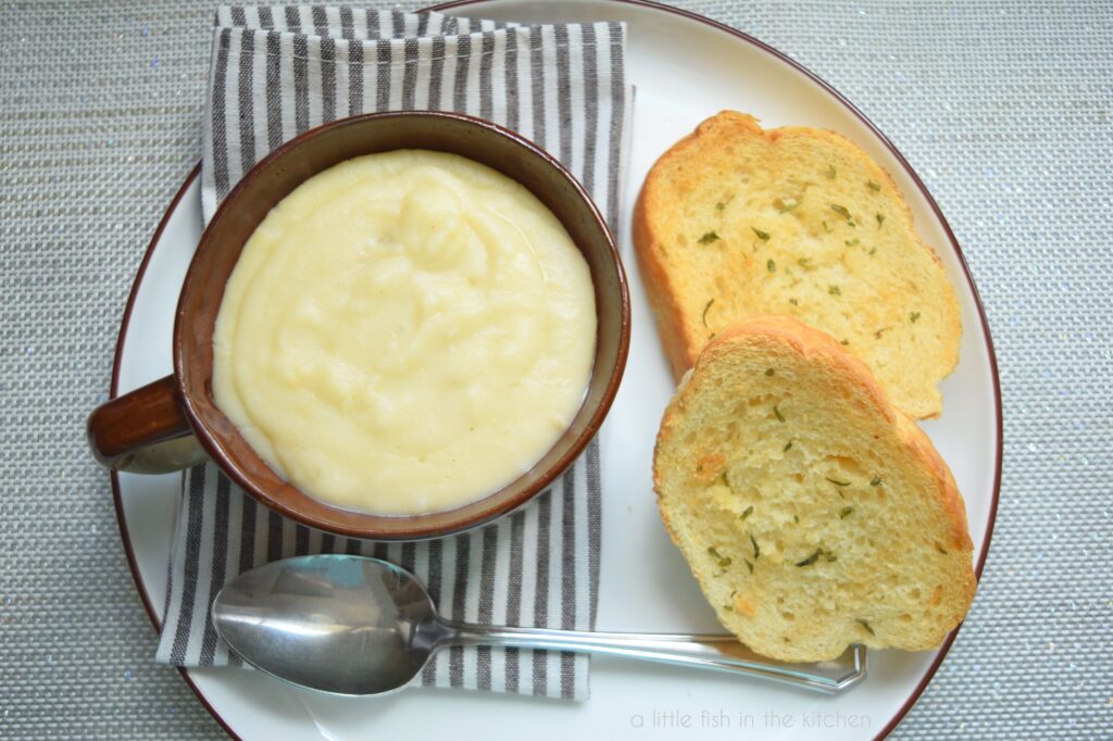 Creamy potato soup is in a brown ceramic bowl on top of a white plate with a brown rim. The bowl sits on top of a brown and white striped cloth napkin. Two pieces of toasted garlic bread are beside the bowl on the plate. A spoon is beside the bowl. 