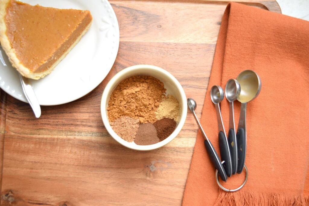 A white ramekin filled with five small piles of warm spices sits on a wooden cutting board. A burnt orange colored cotton tea towel with fringed ends rests just beside the bowl. A set of measuring spoons lies on the towel. On the opposite side of the cutting board, a slice of pumpkin pie is ready to serve on a small white dessert plate.