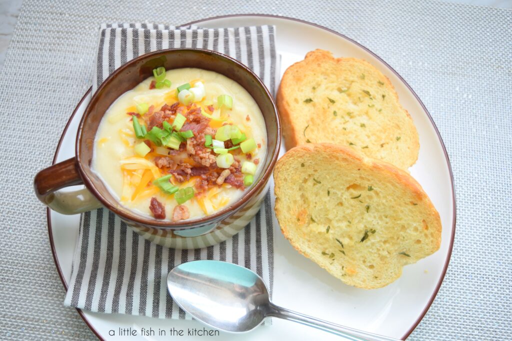 A bowl of crock pot potato soup is topped with shredded cheddar cheese, bacon bits and sliced green onions. It's ready to be served and eaten with two garlic bread slices on the side.