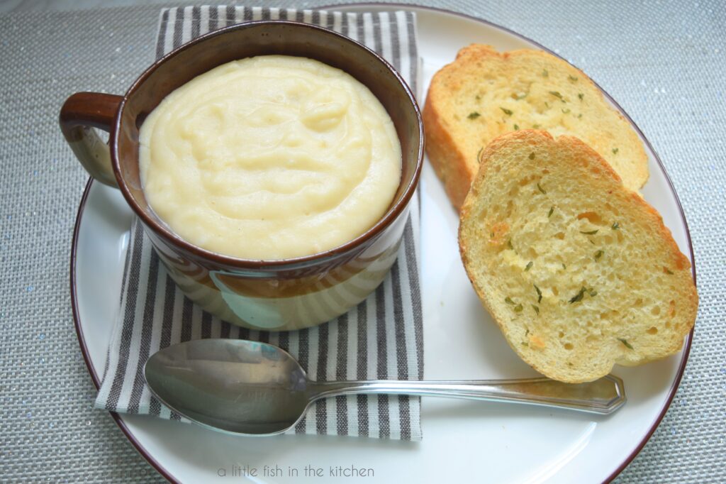 Creamy, golden potato soup is ready to serve in a brown ceramic bowl. The soup is served with two slices of buttery garlic toast and a spoon on the side. 