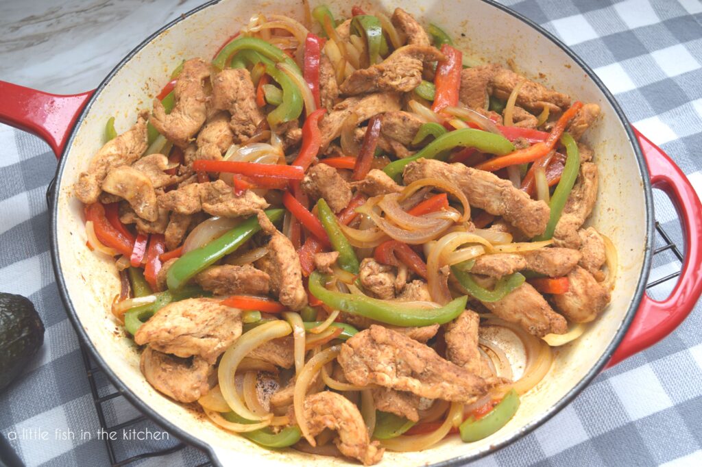 A large, red coated cast iron skillet is filled with sizzling chicken fajitas. The chicken pieces are nestled in with savory, multi-colored peppers and onions. A sliced lime is sitting just beside the hot skillet on a gray and white checker tea towel.