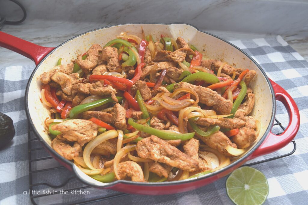 A large, red coated cast iron skillet is filled with sizzling chicken fajitas. The chicken pieces are nestled in with savory, multi-colored peppers and onions. A sliced lime is sitting just beside the hot skillet on a gray and white checker tea towel.
