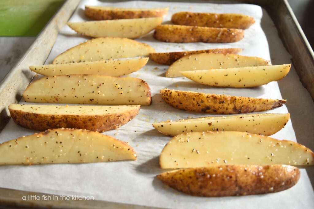 Fresh potato wedges are oiled, seasoned and spread out on a prepared cooking sheet. They are ready for the oven. Specks of ground black pepper and dehydrated garlic are visible on the ivory flesh of the cut potatoes. 