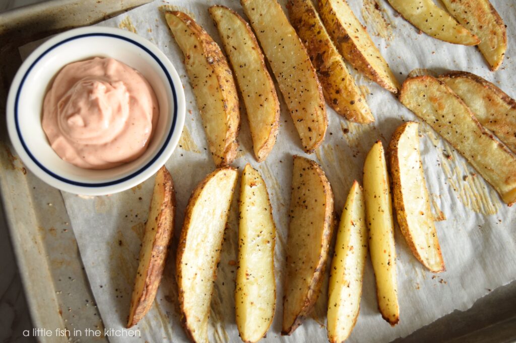 The oven-fried potatoes look golden and crispy laying on a parchment paper-lined baking sheet. There is a small white bowl with a blue rim on the baking sheet with a light red colored french fry dipping sauce. The Pigskin Potatoes are ready to eat!