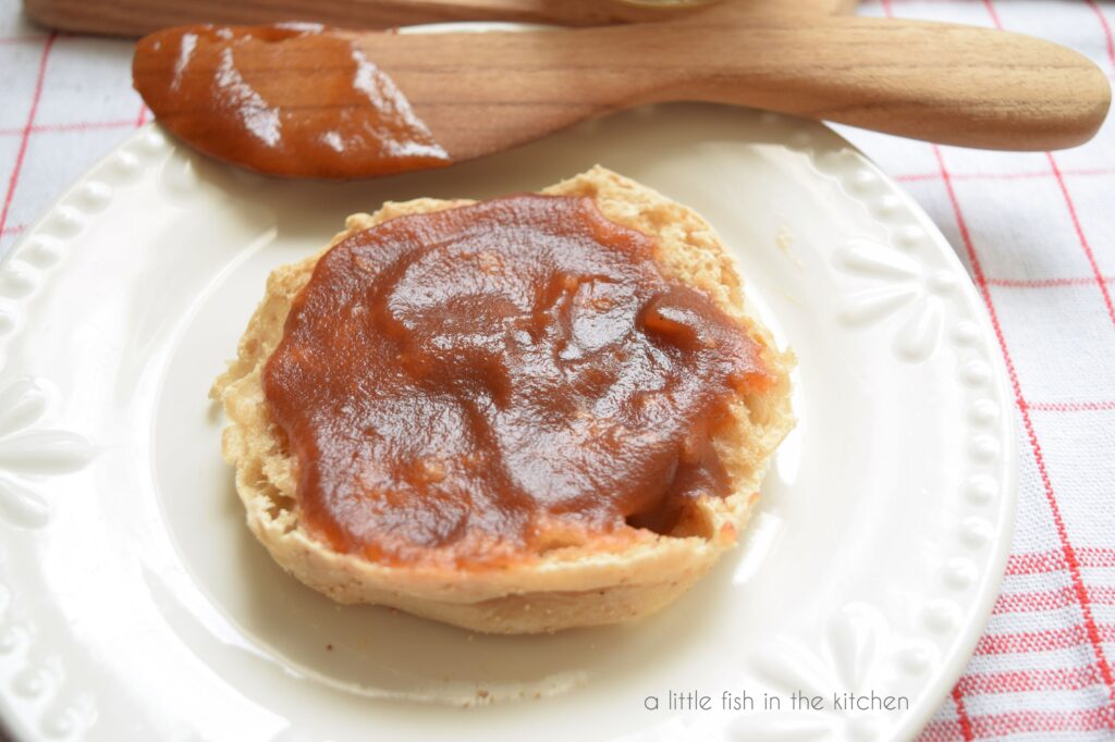 Smooth, heavily spiced, amber apple butter is smeared on a sliced English muffin, ready to enjoy. The English muffin sits on a small white ceramic plate which is on top of a red and white plaid tea towl. A small wooden butter knife still covered in apple butter rests on the plate too. 