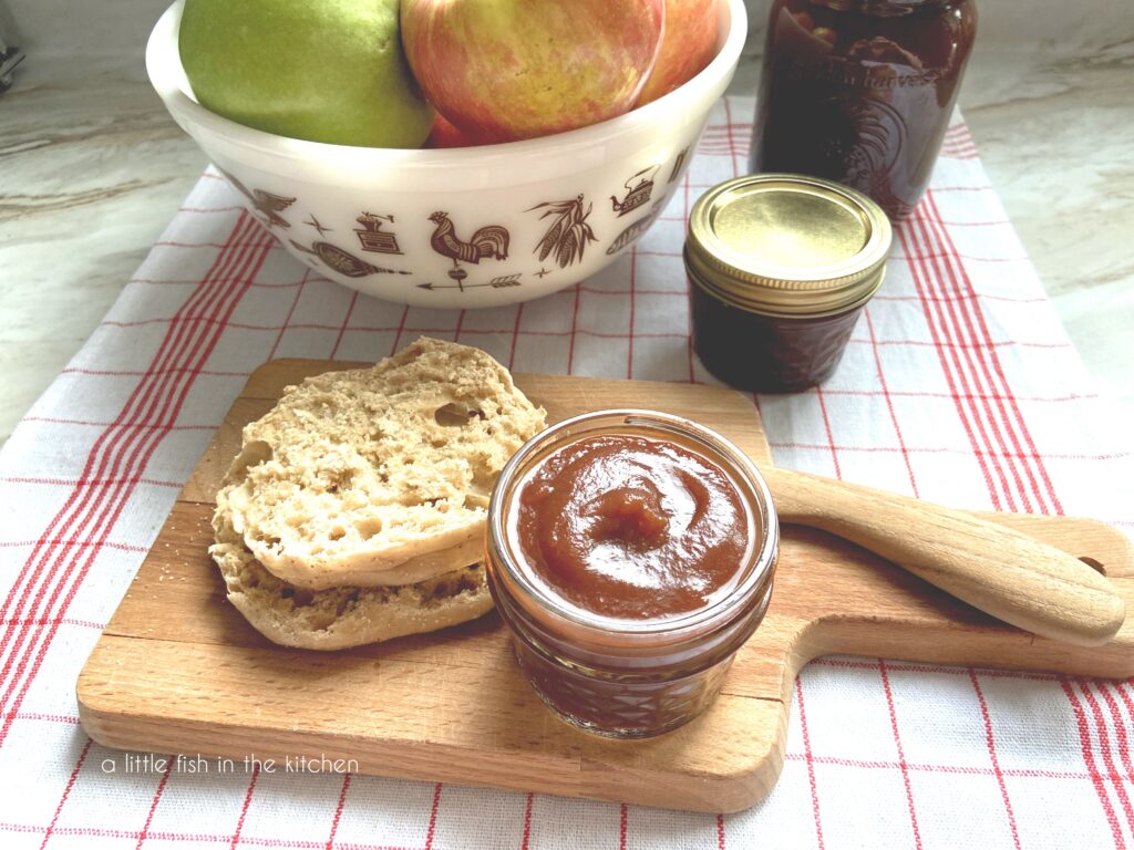 A small glass mason jar filled with golden brown homemade apple butter sits on a wooden cutting board. A sliced, toasted English muffin and a wooden butter knife sit beside beside the jar. 