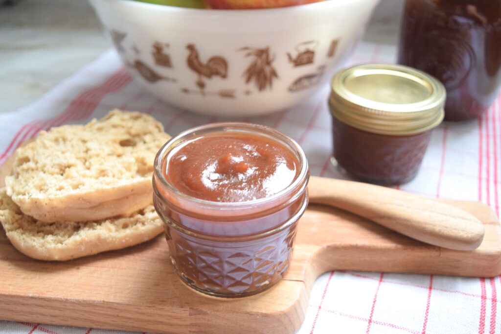 A small glass jar filled with dark-amber colored apple butter sits on a small wooden cutting board. It looks smooth, flavorful and ready to serve on a sliced toasted English muffin. A large vintage bowl filled with colorful fresh apples and another small sealed mason jar filled with dark amber apple butter sit in the background.