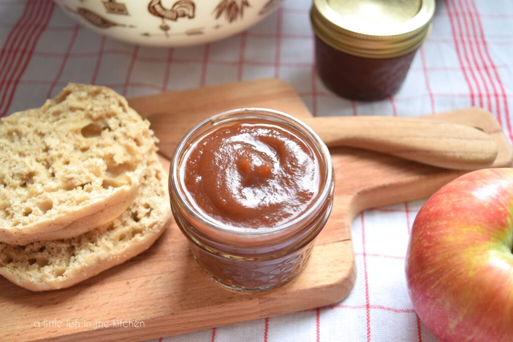 A small glass mason jar filled with golden brown homemade apple butter sits on a wooden cutting board. A sliced, toasted English muffin and a wooden butter knife sit beside beside the jar. 