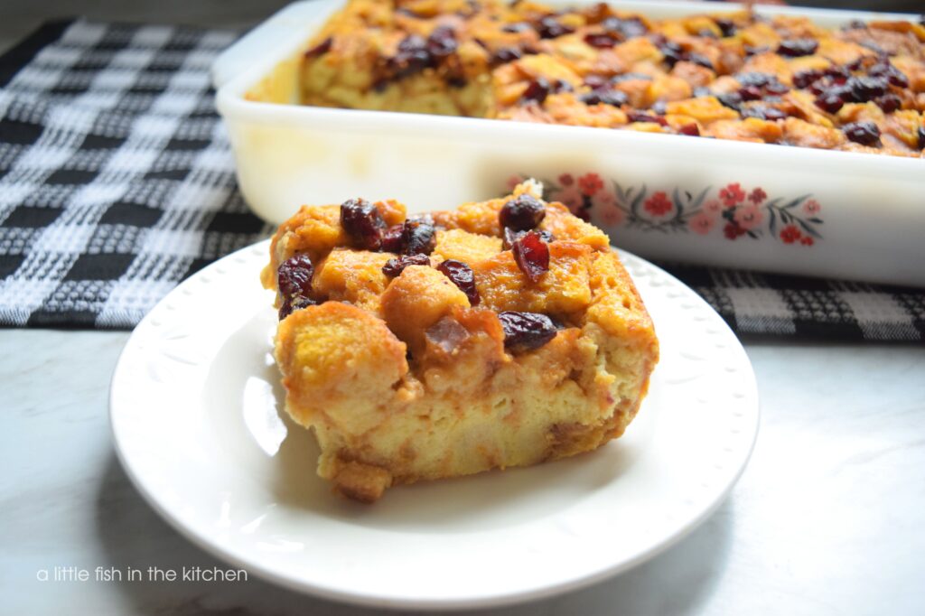 A slice of moist, amber-colored pumpkin bread pudding is sitting on a small white dessert plate. A sprinkling of cranberries are on the top of the dessert. The rest of the dessert is in a white baking dish with a pink, red, and burgandy floral pattern that sits slightly blurred in the background.