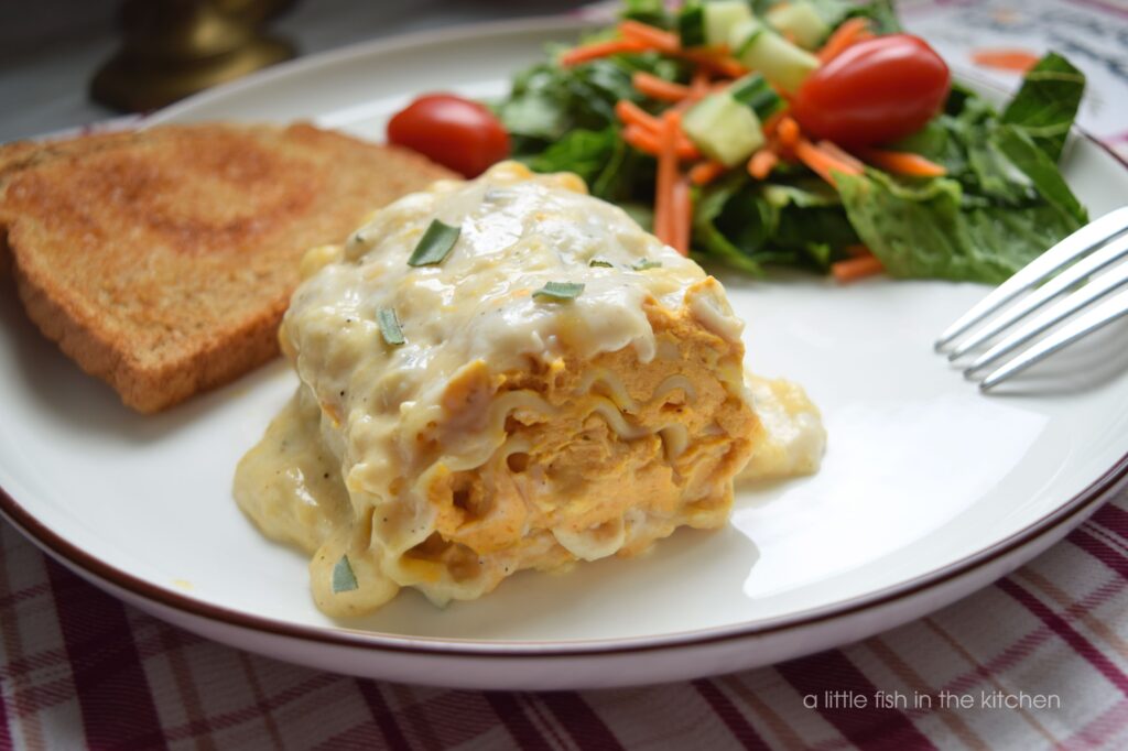 A side view of a pumpkin lasagna roll shows a creamy orange pumpkin and cheese filling and layers of scallopped lasagna noodles. It's topped with a white creamy sauce. A green salad and buttery garlic bread are also on the plate and ready to eat.