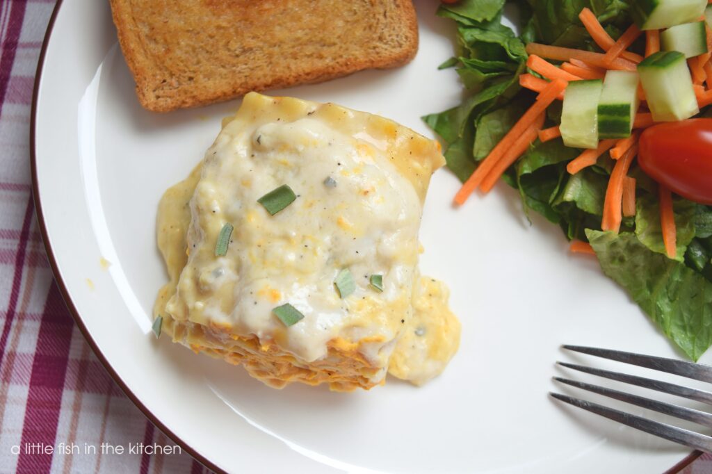 A single pumpkin lasagna roll sits on a white plate. It's covered with a generous pour of creamy bechamel sauce and garnished with minced fresh sage. It's served with a garden salad and a piece of garlic toast. 