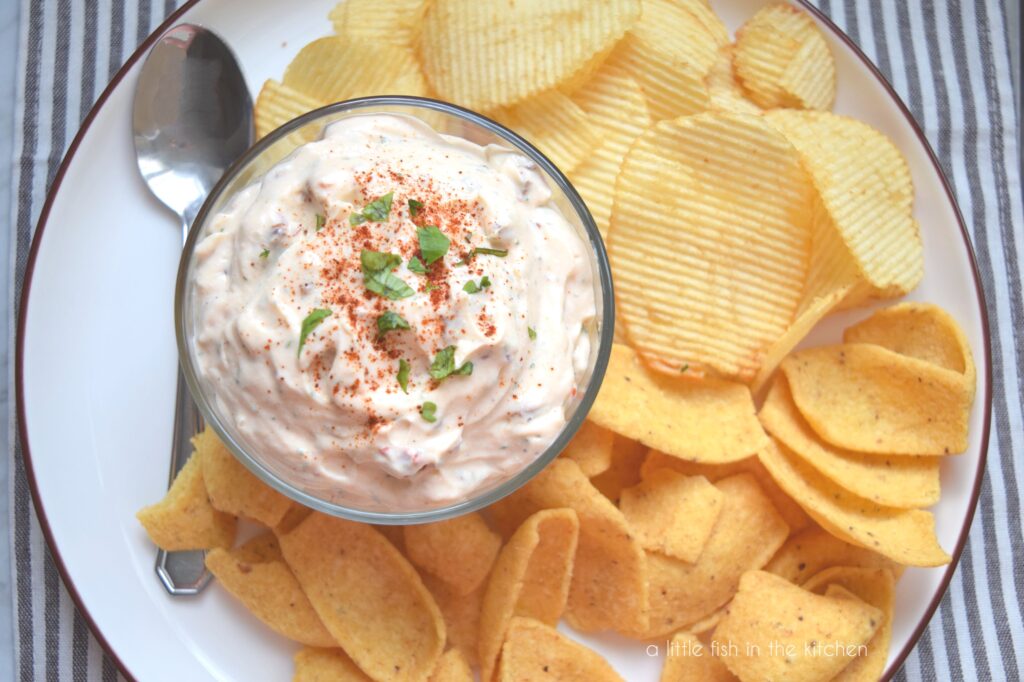 A small clear bowl is filled with a chip dip with a light pink hue, it's garnished on top with a sprinkle of chili powder and finely chopped, fresh green cilantro. The serving plate is filled with corn chips and ridged potato chips, ready to dip!