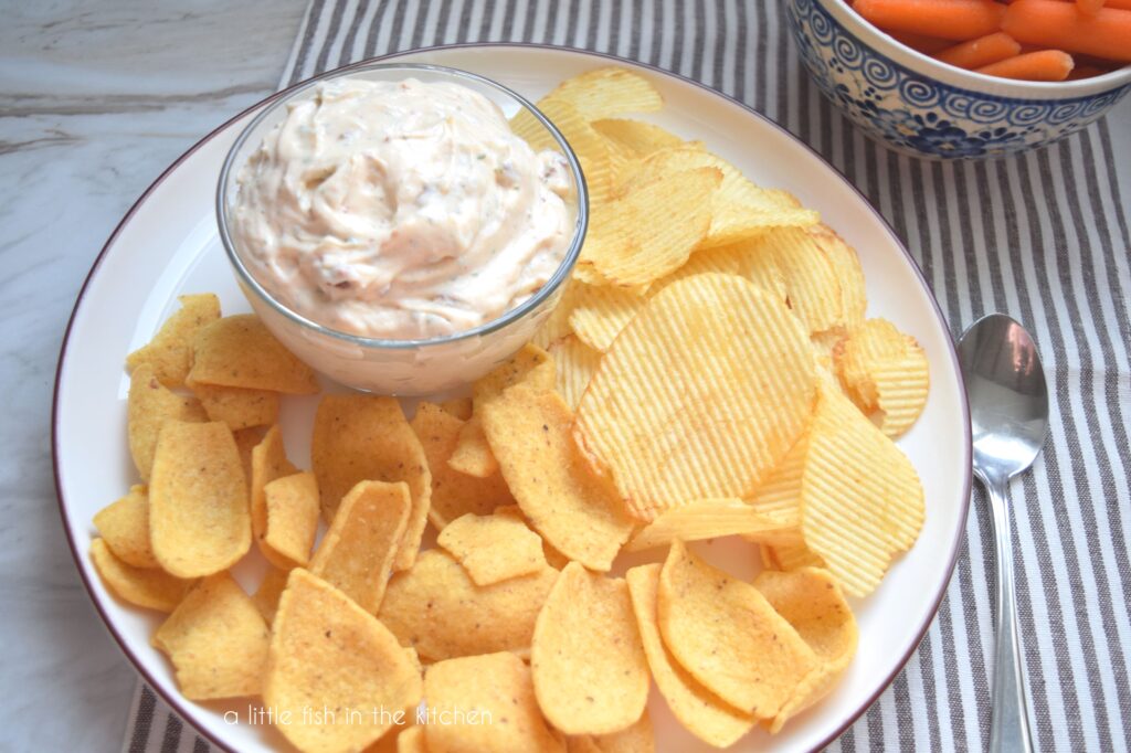 A small clear bowl filled with chipoltle ranch chip dip is on a white serving plate. The small bowl is surrounded by a mixture of corn chips and ridged potato chips. There's small bowl filled with baby carrots just off to the side of the plate. It's ready to enjoy! 