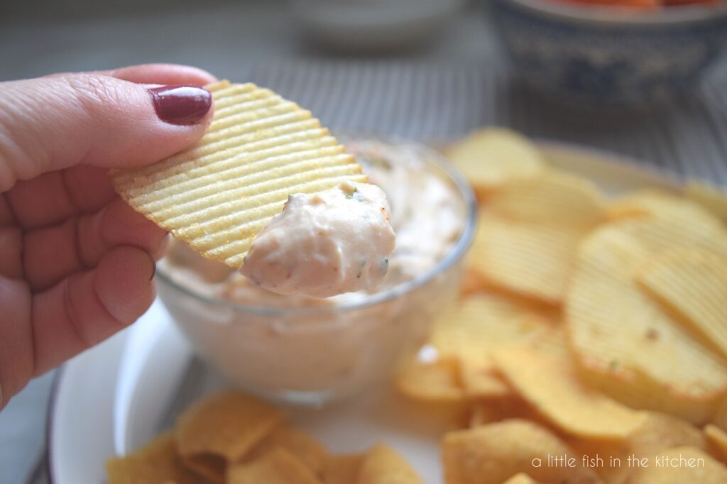 A hand holds a ridged potato chip that has a hearty teaspoon of dip on the end. Spices, and bits of chopped cilantro are visible in the dip. An assortment of chips and corn chips on a platter is slightly blurred in the background. 