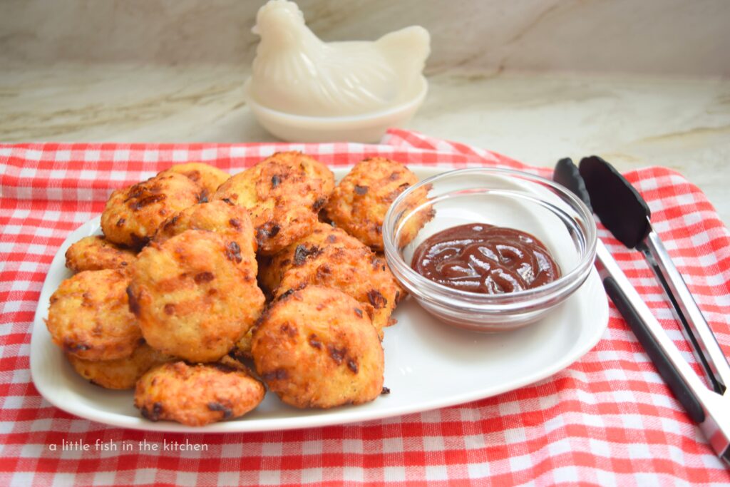 A crispy, golden batch of low carb chicken nuggets is served on a rectangular white plate with a small bowl of barbecue sauce on the sicde. The plate sits on a red and white checkered tea towel and metal tongs are there to serve them. 