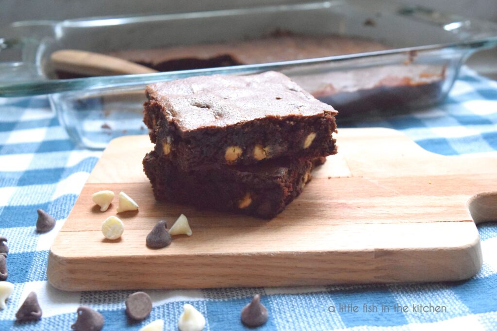 Two brownies are stacked on top of a small wooden cutting board. The crumb of the brownies is a medium brown color and a slightly fudgy texture. White chocolate chips and semi-sweet chocolate chips are seen along the sides of the sliced brownies. The small cutting board sits atop a blue and white checkered tea towel and the glass baking dish with the remainer of the brownies is slightly blurred in the background. 