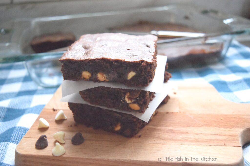 Three scratch-baked triple chocolate brownies are stacked on top of a small wooden cutting board with squares of parchment paper layered between them. Bits of sweet white and semi-sweet chocolate chips are seen on the sides of the dark brownie crumb. They are ready to be served or packed up to share. 