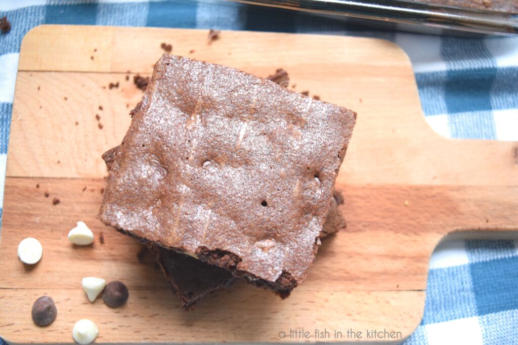 The crackly tops of these homemade brownies look very inviting. The brownies are sitting on a small wooden cutting board. The cutting board is on top of a blue and white checkered tea towel. Whole white chocolate chips and semi-sweet chocolate chips are scattered on the cutting board next to the stacked brownies for a decorative effect.