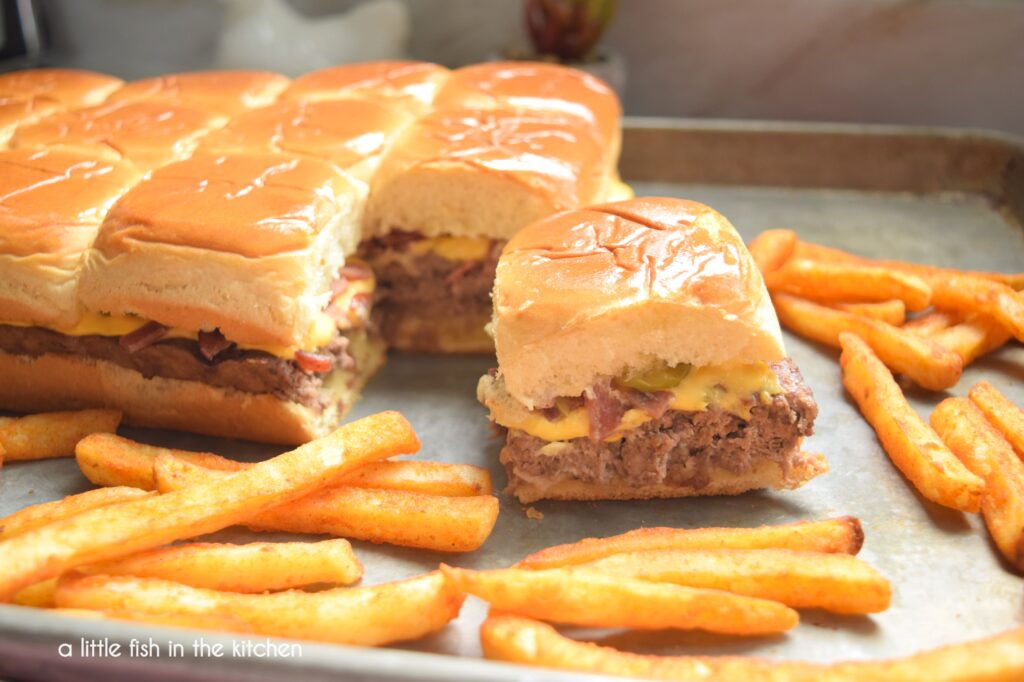 A single bacon cheeseburger slider is pulling away from a set of oven-baked sliders on Hawaiian rolls. A seasoned ground beef patty, melted  American cheese, pickles and bacon bits are all visibly sandwiches between the Hawaiian rolls. Crisp French fries are scattered around the sliders for serving on the sheet pan. 