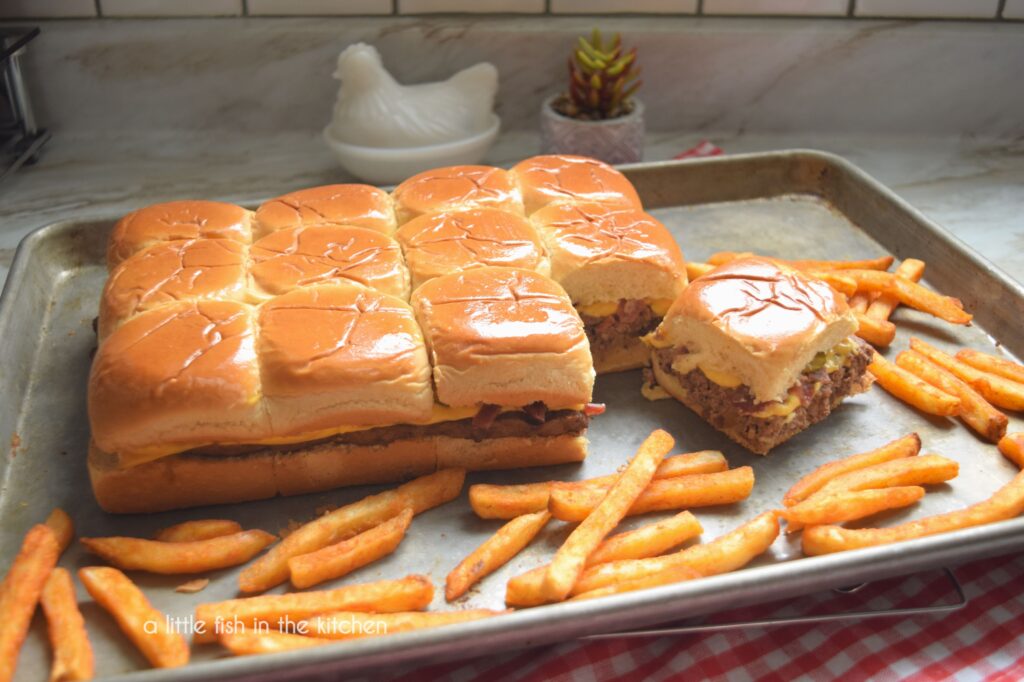 Nine freshly-prepard, flavorful cheeseburger sliders are served on a metal baking sheet. The savory baked sliders are made with buttery Hawaiian rolls, and the meat patty, cheese, pickles and onions are visible from the side of the set of sliders. Seasoned French fries are scattered around the sheet pan as a grab and go side option. 