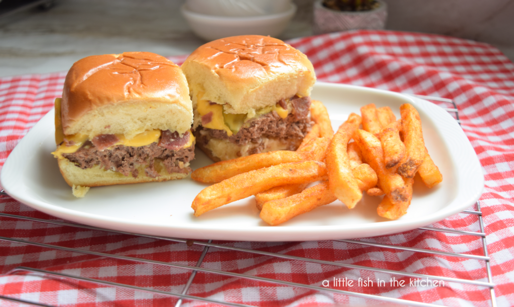 Two cheesy, buttery bacon cheeseburger sliders are served with crispy seasoned French fries on a rectangular white plate. The plate is served on red and white checkered tea towel. .