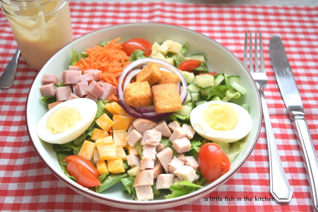 A colorful chef salad is served in a large, ceramic white salad bowl with a brown rim. The bowl is set atop a red and white checkered tablecloth. A fork and knife are set beside the bowl. Green, torn lettuce is topped with a rainbow of chopped, raw veggies including carrots, cucumber, red onion slices, cherry tomatoes. Diced deli ham and smoked turkey, as well as a sliced hard cooked egg are included in this hearty dinner salad.